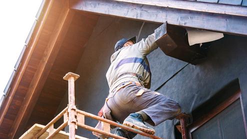 Arbeiter steht auf einem Gerüst und befestigt ein Element unter dem Dach eines Hauses.